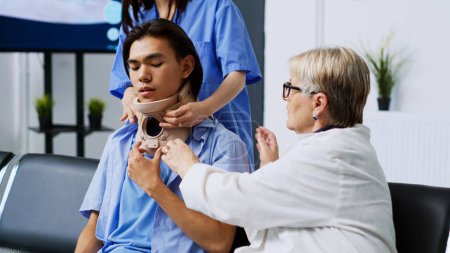 Téléchargez les photos : Traumatologue aidant le patient asiatique à prendre un collier cervical dans la salle d'attente de l'hôpital, expliquant le traitement médical. Médecin âgé recevant un soutien médical lors de la visite de contrôle rendez-vous - en image libre de droit