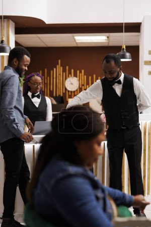 Téléchargez les photos : Jeune afro-américain souriant gars groom fournissant un service à la clientèle aux clients de l'hôtel, déplacer ses bagages, valise de transport à la chambre après l'enregistrement. Bellhop et la communication touristique à la réception - en image libre de droit