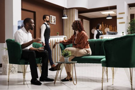 Téléchargez les photos : Les Afro-Américains attendent l'enregistrement de la chambre, assis dans le salon de l'hôtel de luxe. Couple joyeux arrivant à la station balnéaire chère moderne, se préparant pour l'inscription à la réception. - en image libre de droit