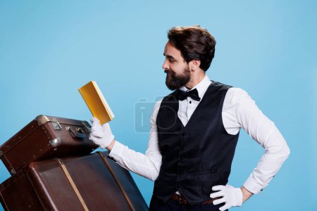 Hotel concierge reading book in studio posing dressed in formal attire, having fun with literature or fiction tales. Young bellboy employee lecturing novel story, intelligent doorkeeper.