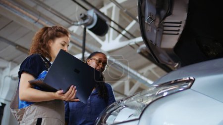 Photo for Licensed technician in garage using laptop to follow checklist while doing maintenance on vehicle, talking with client. Worker in car service does checkup on automobile, ground level shot - Royalty Free Image