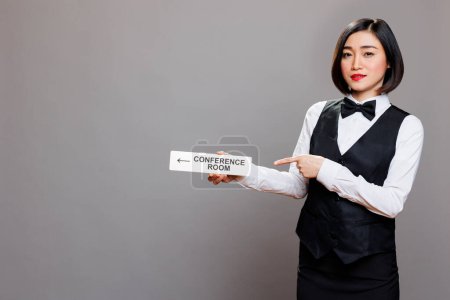 Photo for Asian woman receptionist holding conference room directional tabletop signage and pointing with finger portrait. Hotel employee in uniform showing boardroom steel sign and looking at camera - Royalty Free Image