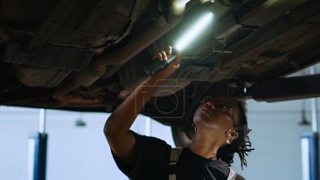 Photo for Licensed mechanic standing underneath suspended car in garage workspace, using work light to check for damages and tablet to order new replacing parts after finding issues during annual maintenance - Royalty Free Image