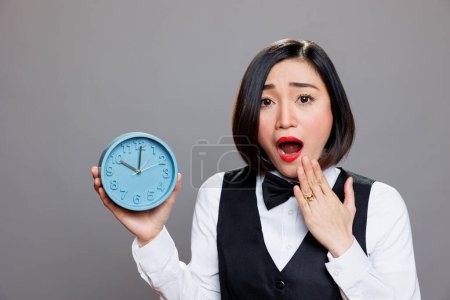 Photo for Shocked overslept asian waitress holding alarm clock and looking at camera while covering open mouth. Young anxious woman receptionist showing time while running late portrait - Royalty Free Image