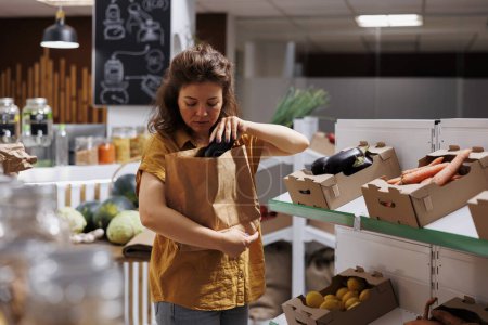 Téléchargez les photos : Femme dans un magasin zéro déchet achetant des légumes naturels cultivés à la ferme, cueillant des aubergines mûres. Client dans une épicerie locale gratuite en plastique cherchant à acheter des aliments sains, en utilisant un sac en papier biodégradable - en image libre de droit