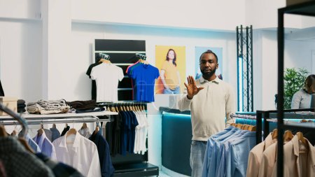 Téléchargez les photos : Acheteur afro-américain utilisant l'hologramme en boutique pour vérifier les nouvelles marchandises de magasin de vêtements avec la réalité augmentée. Client masculin regardant des séquences holographiques, intelligence artificielle. - en image libre de droit