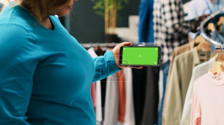 Foto de Mujer afroamericana analizando el teléfono con plantilla de pantalla verde, mirando la pantalla aislada en la boutique moderna. Elegante cliente de compras de ropa de moda en la tienda de ropa. Concepto de moda - Imagen libre de derechos