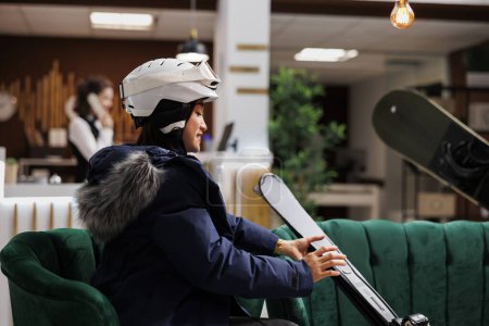 Téléchargez les photos : Femme asiatique en veste d'hiver confortablement assise sur le canapé avec son équipement de ski à la réception de l'hôtel. Femme enthousiaste avec casque de neige plaçant ses skis à proximité dans le salon. - en image libre de droit
