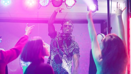 Téléchargez les photos : DJ afro-américain mélangeant audio sur scène dans une boîte de nuit, homme cool faisant la fête avec un groupe de personnes sur la piste de danse. Funky adulte créant des sons de mélange électronique à la station de panneau, clubbing. Trépied. - en image libre de droit