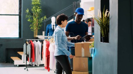 Photo for Manager helping courier with packages, carrying cardboard boxes in modern boutique. African american deliveryman explaining shipping details to worker in clothing store. Fashion concept - Royalty Free Image