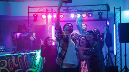 Photo for Diverse group of adults taking photos on dance floor under colorful spotlights, having fun at party celebration. Cheerful men and women enjoying disco event filming videos. Handheld shot. - Royalty Free Image