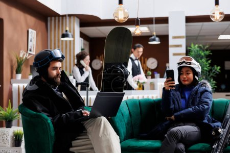 Photo for Boyfriend using laptop and girlfriend with mobile phone enjoying digital communication. Wintersports enthusiasts in hotel lobby for ski and snowboarding fun waiting with their smart devices. - Royalty Free Image