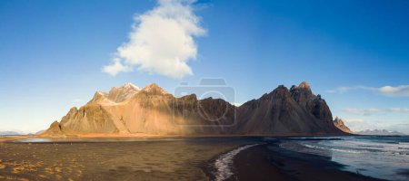 Foto de Vista panorámica de las colinas rocosas de vestrahorn en la península icelándica, con una playa de arena negra única y un entorno nórdico al aire libre. Increíble paisaje creado por la cima de la montaña y las olas del océano. - Imagen libre de derechos