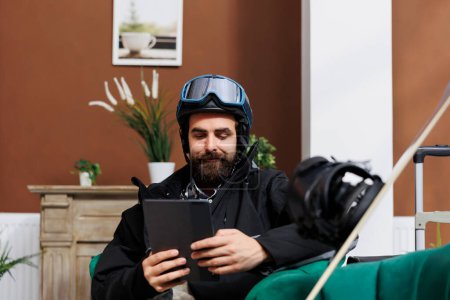 Photo for Male guest seated in cozy hotel lobby surfing the net on smart tablet. Caucasian man with device dressed in snow gear and ski goggles resting on forehead hints at upcoming adventure on winter slopes. - Royalty Free Image