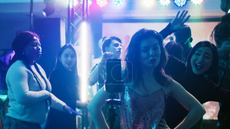 Young adult filming video at party, using mobile phone to record vlog of group of people having fun at club. Smiling person enjoying night out partying with friends, entertainment.