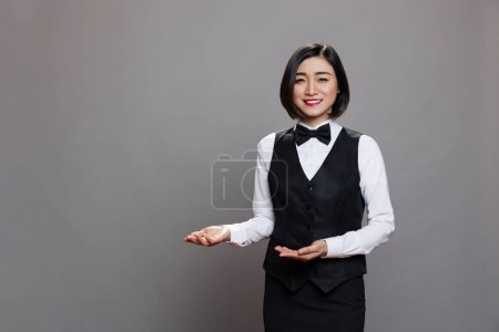 Téléchargez les photos : Serveuse asiatique souriante portant un uniforme de restaurant montrant avec les mains et saluant le portrait des invités. Réceptionnistes professionnels accueillant les clients et regardant la caméra - en image libre de droit