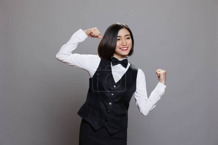 Photo for Smiling cheerful attractive asian waitress laughing while showing bicep. Joyful young woman receptionist wearing professional uniform flexing hands muscles while posing in studio - Royalty Free Image