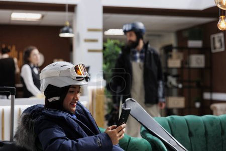 Photo for Eager asian woman waits in ski hotel reception, using her cellphone while anticipating winter vacation booking process. Tourist dressed in snow gear occupies lounge area, engrossed in her mobile - Royalty Free Image