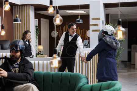 Asian bellboy with warm smile extends a welcoming hand to female visitor, ensuring personalized and enjoyable stay. Male traveler waits on sofa for reservation while young woman in snow gear arrives.