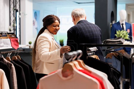 Foto de Amistosa sonriente asistente de tienda de mujer negra ayudando a los clientes mayores a elegir la ropa en la boutique de moda. Útil vendedora afroamericana agradable sirviendo ayudando al comprador en la tienda de ropa - Imagen libre de derechos