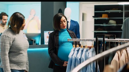 Foto de Mujer embarazada asiática revisando ropa moderna en bastidores, futura madre que busca comprar ropa formal. Joven cliente con el vientre del embarazo examinando mercancía tienda de moda, baby bump. - Imagen libre de derechos