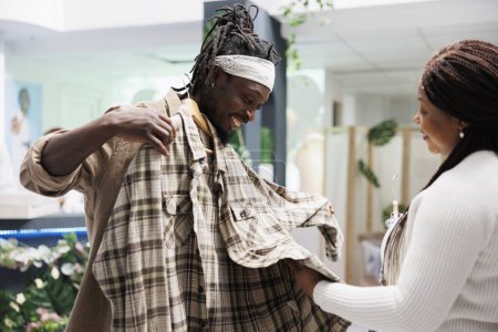 Foto de Hombre afroamericano sosteniendo camisa a cuadros al cuerpo, mostrando a su esposa y pidiendo consejo en la tienda de ropa. Pareja de compras y probarse ropa casual en la boutique de moda - Imagen libre de derechos