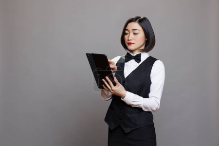 Foto de Joven camarera asiática en uniforme de control de menú de café en la tableta digital. Personal de servicio de catering administrando pedidos en línea y tocando en la pantalla táctil mientras posan en el estudio sobre fondo gris - Imagen libre de derechos
