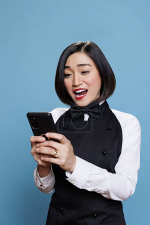 Photo for Excited cheerful asian woman dressed in waitress uniform texting online on smartphone. Young attractive receptionist typing message on mobile phone while posing in studio - Royalty Free Image