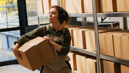 Photo for Supervisor organizing boxes on racks, carrying packages before preparing orders for retail shipment. Young adult listening music checking merchandise stock in storage room. Handheld shot. - Royalty Free Image
