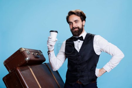 Téléchargez les photos : Elégant concierge d'hôtel avec boisson posant sur fond bleu en studio, agissant en toute confiance en tant que groom ou portier. Employé élégant sert une tasse de thé de café. - en image libre de droit