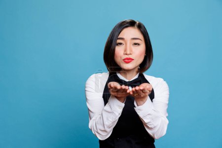 Photo for Smiling attractive asian waitress wearing uniform sending air kiss and looking at camera. Beautiful young woman receptionist expressing love and affection portrait in studio - Royalty Free Image