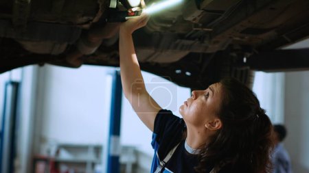 Photo for Woman working on suspended car in garage, checking parts during annual checkup. Professional in repair shop workplace walking underneath vehicle, scanning it using work light - Royalty Free Image