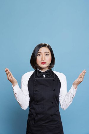 Foto de Restaurante joven mujer asiática empleada rezando con los brazos abiertos y mirando hacia arriba. Café atractiva camarera con uniforme profesional meditando, practicando la relajación en el estudio - Imagen libre de derechos