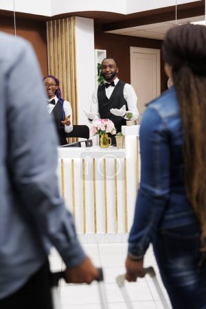 Téléchargez les photos : Personnel de réception afro-américain accueillant accueillant les clients avec le sourire à l'hôtel. Vue arrière du couple noir tirant des valises allant à la réception pour confirmer la réservation - en image libre de droit