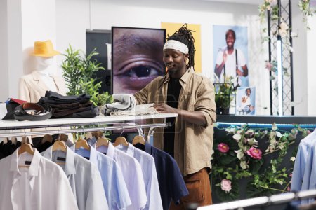 Foto de Sonriente joven afroamericano hombre elegir ropa casual en la tienda de ropa centro comercial. Cliente de pie cerca de rack con camisas y ropa de búsqueda mientras revisa el tamaño - Imagen libre de derechos