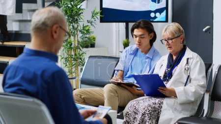 Photo for Elderly doctor helping asian patient to fill medical report, explaining disease expertise. Medic discussing treatment with young adult during checkup visit in hospital waiting area. Medicine service - Royalty Free Image