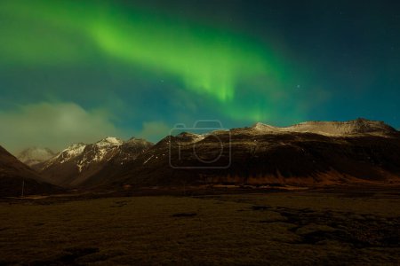 Foto de Paisaje islandés con luces boreales que cubren las cumbres nevadas de las montañas, fotografía nocturna. Aurora boreal nórdica que ilumina el cielo estrellado con verde y azul, paisaje con panorama cautivador. - Imagen libre de derechos