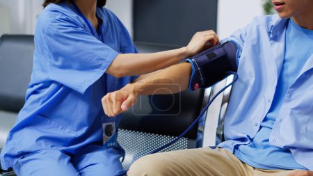 Foto de Enfermera asiática revisando la hipertensión del paciente usando tonómetro médico durante la consulta de revisión en la sala de espera del hospital. Asistente examinando la presión arterial del hombre mayor en recepción - Imagen libre de derechos