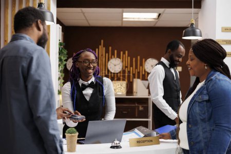Photo for Smiling positive African American woman front desk agent receiving contactless payment from guests at reception counter. Black couple of tourists paying for room with credit card at check-in - Royalty Free Image