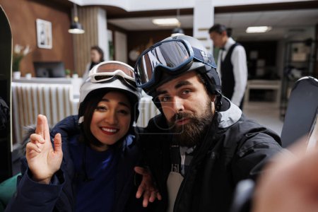 Téléchargez les photos : Jeunes invités capturant des aventures hivernales avec des selfies sur smartphone. Mélange de technologie et de vacances en hiver pour couple en vêtements de neige lunettes de ski et casque dans une station de montagne exclusive. - en image libre de droit