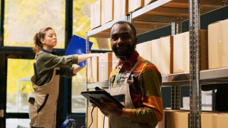 Téléchargez les photos : Un Afro-Américain utilisant un scanner dans un entrepôt pour scanner des codes-barres sur des produits stockés dans des boîtes. Employé masculin examinant des marchandises de fret sur des supports et des étagères, examinant la logistique. - en image libre de droit