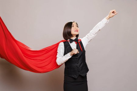 Photo for Asian waitress wearing superwoman cloak raising clenched fist and looking upwards. Catering service woman employee flying in fluttering hero red cape while posing in studio - Royalty Free Image