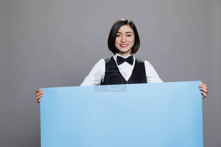 Photo for Smiling asian waitress holding blank banner with copy space for restaurant advertising and looking at camera. Cheerful woman receptionist in uniform standing with empty whiteboard in hands portrait - Royalty Free Image