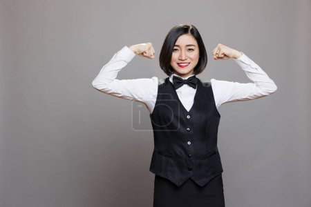 Photo for Cheerful attractive strong asian woman receptionist showcasing bicep muscles portrait. Restaurant waitress in uniform showing power and strength concept while looking at camera - Royalty Free Image