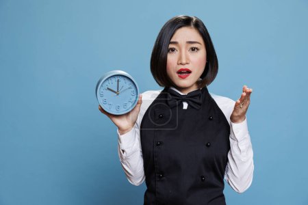 Photo for Asian waitress holding alarm clock with surprised facial expression portrait. Worried young woman cafe employee wearing professional uniform showing time and looking at camera - Royalty Free Image