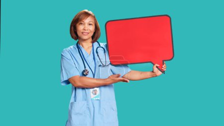 Téléchargez les photos : Heureuse infirmière asiatique souriante tenant bulle rouge signe de l'espace vide pour le texte. Spécialiste de la santé souriant présentant le carton à bulles de pensée, isolé sur fond bleu studio - en image libre de droit