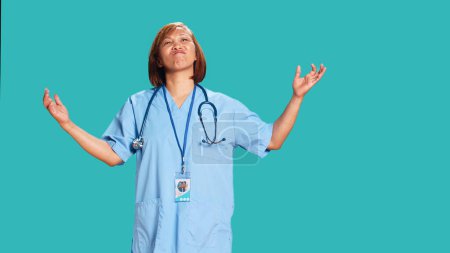 Photo for Confused asian nurse frustrated about not knowing answer to patient inquiry while at work. Stumped healthcare specialist doing perplexed gesturing, isolated over blue studio background - Royalty Free Image