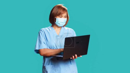 Photo for Happy medical nurse chatting online with patient on laptop during telemedicine checkup appointment. Joyous asian woman wearing protective face mask using gadget, isolated over blue studio background - Royalty Free Image