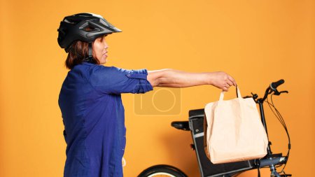 Photo for BIPOC food delivery worker reaching order address, waiting for customer to answer door. Courier woman greeting client, offering takeaway lunch bag, isolated over orange studio background - Royalty Free Image