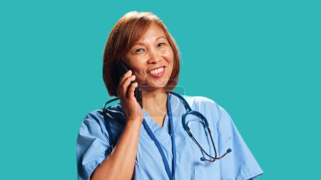 Photo for Upbeat BIPOC clinic employee wearing protective uniform having lively chat while on call with friend. Close up shot of nurse happily answering phone while at work, isolated over studio background - Royalty Free Image